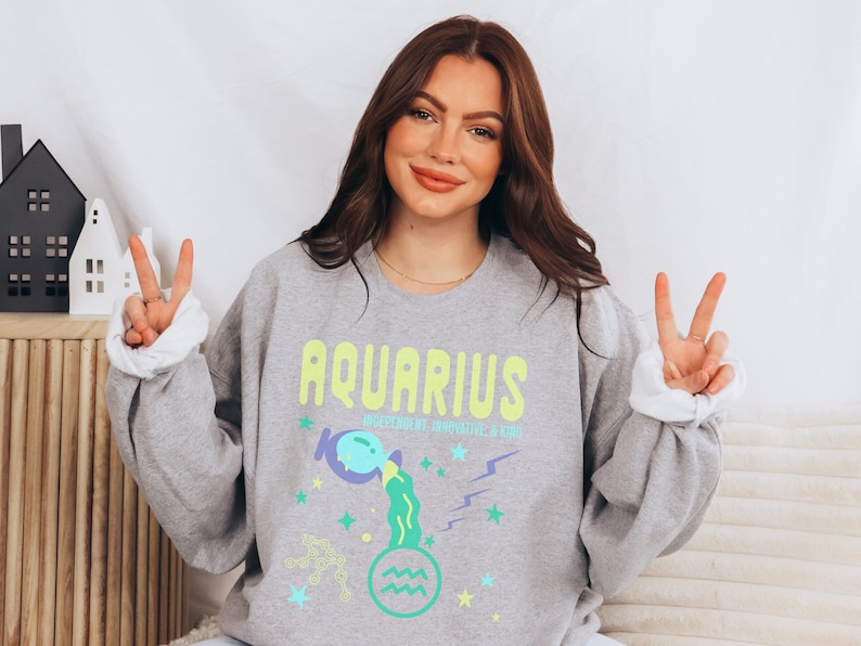K&ouml;nnte beinhalten: Grauer Pullover mit einem grafischen Design des Sternzeichens Wassermann. Das Design zeigt einen blauen und gr&uuml;nen Wasserkrug, Sterne und den Text "Wassermann, Unabh&auml;ngig, Innovativ und Freundlich".