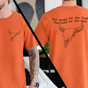 May include: Orange t-shirt with a bull skull graphic on the front and the text "Too Soupy for the Trash Too Solid for the Sink" on the back.