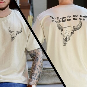 May include: A beige t-shirt with a skull graphic on the front and the text "Too Soupy for the Trash Too Solid for the Sink" on the back.