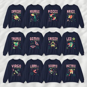 May include: Twelve navy blue sweatshirts with zodiac signs and names printed on the front. The signs are illustrated in pink, green, and yellow. The signs are: Capricorn, Aquarius, Pisces, Aries, Taurus, Gemini, Cancer, Leo, Virgo, Libra, Scorpio, and Sagittarius.