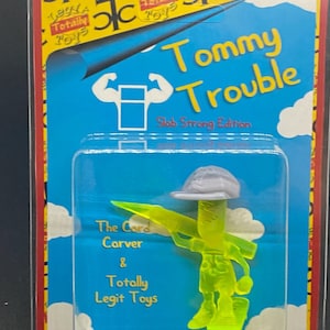 Może przedstawiać: Zielona plastikowa figurka postaci o imieniu Tommy Trouble, marki Totally Legit Toys. Figurka trzyma zielone narzędzie do rzeźbienia i ma na sobie szary kapelusz. Opakowanie jest niebieskie i żółte z napisem "Tommy Trouble Slob Strong Edition".