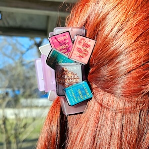 May include: A purple hair clip with a stack of miniature book covers featuring the titles "A Court of Thorns and Roses", "A Court of Wings and Ruin", "A Court of Silver Flames", and "A Court of Frost and Starlight".