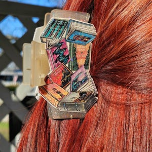 May include: A translucent, glittery hair claw clip featuring a decorative arrangement of miniature book illustrations. The books display titles such as Crown of Midnight, Heir of Fire, Queen of Shadows, and Empire of Storms by Sarah J. Maas.