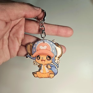 May include: A cartoon keychain featuring a character with a blue and pink hat, blue scarf, and yellow outfit. The keychain is attached to a silver metal ring. The character has large eyes and is smiling. The keychain is held by a hand.