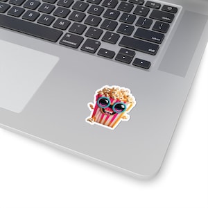 May include: A sticker featuring a cartoon popcorn bucket with a smiling face and sunglasses. The popcorn bucket has pink and white stripes, and the sunglasses are light blue. The sticker is on a laptop.