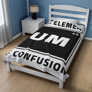 May include: A white and black throw blanket with the text "Element 42 Um Confusion" printed on it. The blanket is on a white bed frame.