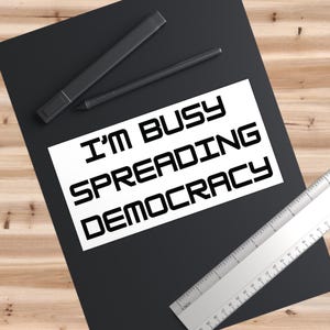 May include: A white sticker with black text that reads "I'm busy spreading democracy". The sticker is on a black surface with a ruler and two black markers.
