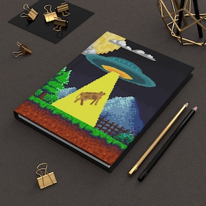 Könnte beinhalten: Ein schwarzes Notizbuch mit einem pixeligen Design einer Kuh, die von einem UFO entführt wird. Das Notizbuch hat einen gelben Lichtstrahl, der vom UFO nach unten strahlt.