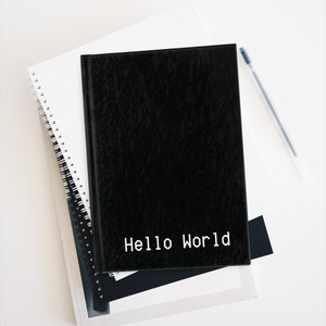 Puede incluir: Una libreta negra con el texto "Hello World" impreso en blanco en la portada. La libreta está encima de una pila de otras libretas y un bolígrafo.