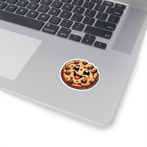 May include: A round sticker featuring a cartoon cookie with a smiling face and chocolate chips. The cookie has a light brown color with darker brown chips and a white border. The sticker is placed on a laptop.