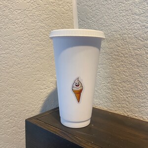 May include: White reusable cup with a lid and a straw. The cup has a sticker of a smiling ice cream cone.