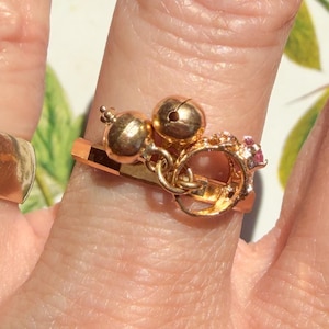 May include: A gold-toned ring featuring two small spherical charms and a decorative element with a tiny pink stone. The ring band is flat and metallic, worn alongside a plain, wide, polished gold-toned band on the adjacent finger.