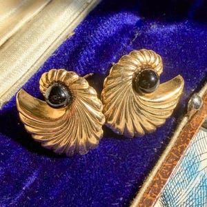 May include: A pair of gold-toned earrings shaped like stylized seashells, each featuring a small, smooth black bead at the center. The earrings have a ribbed, fan-like texture and are displayed inside a rectangular jewelry box with blue velvet.