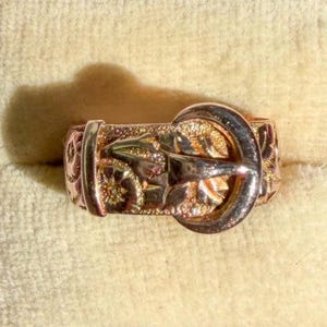 May include: A gold ring designed to resemble a miniature belt with a buckle, featuring an engraved pattern along the band. The ring is presented inside a small, square, red jewelry box with a cream-colored velvet interior lining.