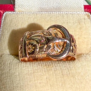 May include: A gold ring designed to resemble a belt with a buckle, featuring intricate engraved floral patterns on the band. The ring is presented inside a red velvet-lined jewelry box with the text "Bishopsgate, LONDON" visible on the interior.