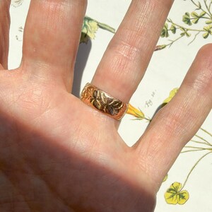 May include: A gold band ring featuring an embossed floral pattern across its surface. The ring has a polished finish and a wide, flat profile, displaying intricate botanical engravings that wrap around the entire circumference of the metal band.
