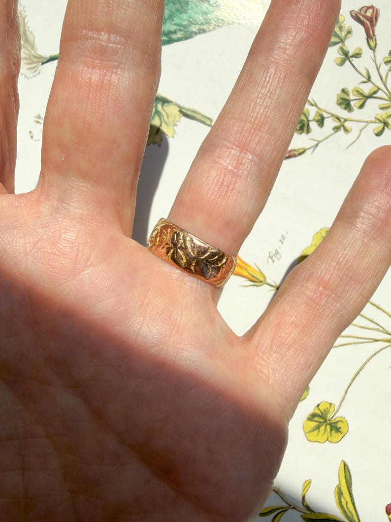 May include: A gold band ring featuring an embossed floral pattern across its surface. The ring has a polished finish and a wide, flat profile, displaying intricate botanical engravings that wrap around the entire circumference of the metal band.