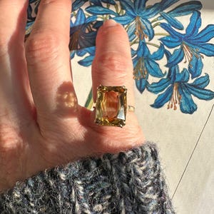 May include: A gold ring with a large, rectangular, amber-colored gemstone. The ring is set on a finger, with a blue floral illustration in the background. The person is wearing a gray sweater.