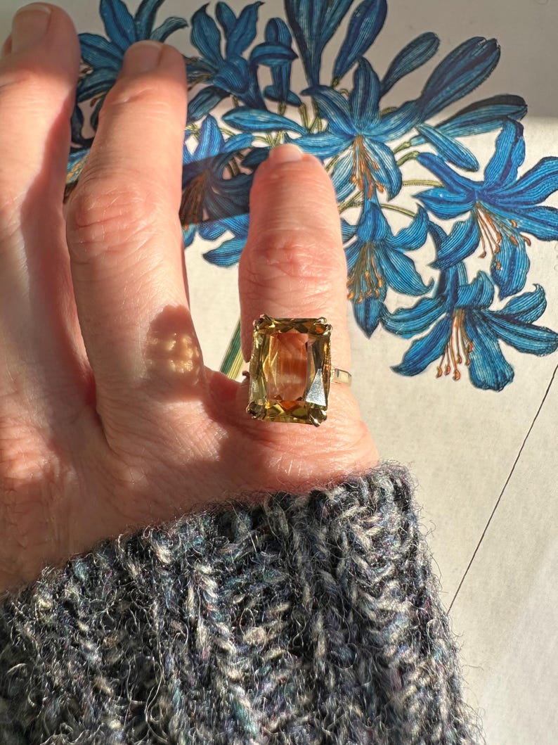 May include: A gold ring with a large, rectangular, amber-colored gemstone. The ring is set on a finger, with a blue floral illustration in the background. The person is wearing a gray sweater.