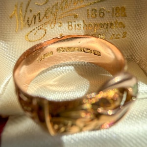 May include: A rose gold ring featuring a buckle design with an engraved pattern on the outer band. The interior of the band displays clear hallmarks including the number 375, indicating gold purity, alongside other small stamped maker marks.