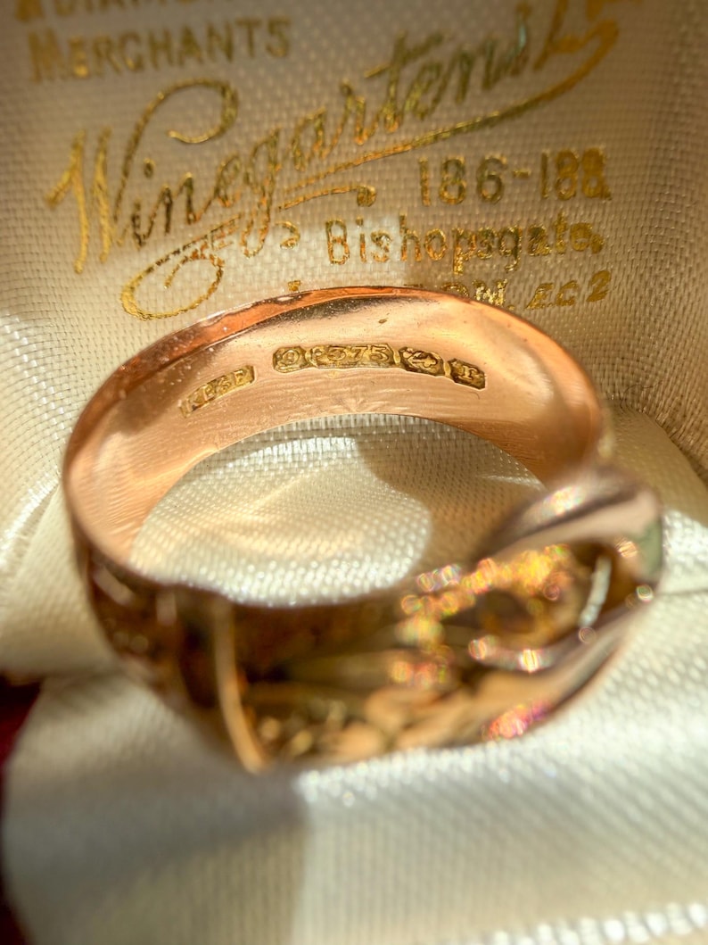 May include: A rose gold ring featuring a buckle design with an engraved pattern on the outer band. The interior of the band displays clear hallmarks including the number 375, indicating gold purity, alongside other small stamped maker marks.