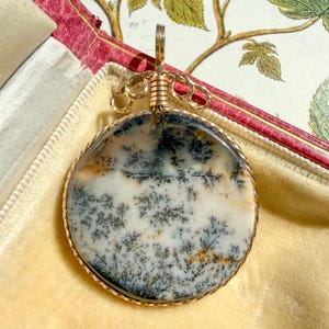 May include: A round dendritic agate pendant featuring a white base with dark, fern-like inclusions and subtle orange accents. The stone is encased in a twisted gold-tone metal wire frame with a decorative wire-wrapped bail at the top.