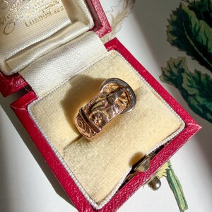 May include: A gold ring shaped like a belt buckle with engraved floral patterns and a textured finish, presented inside a red rectangular jewelry box with a cream-colored velvet interior lining and gold text printed on the inner lid.