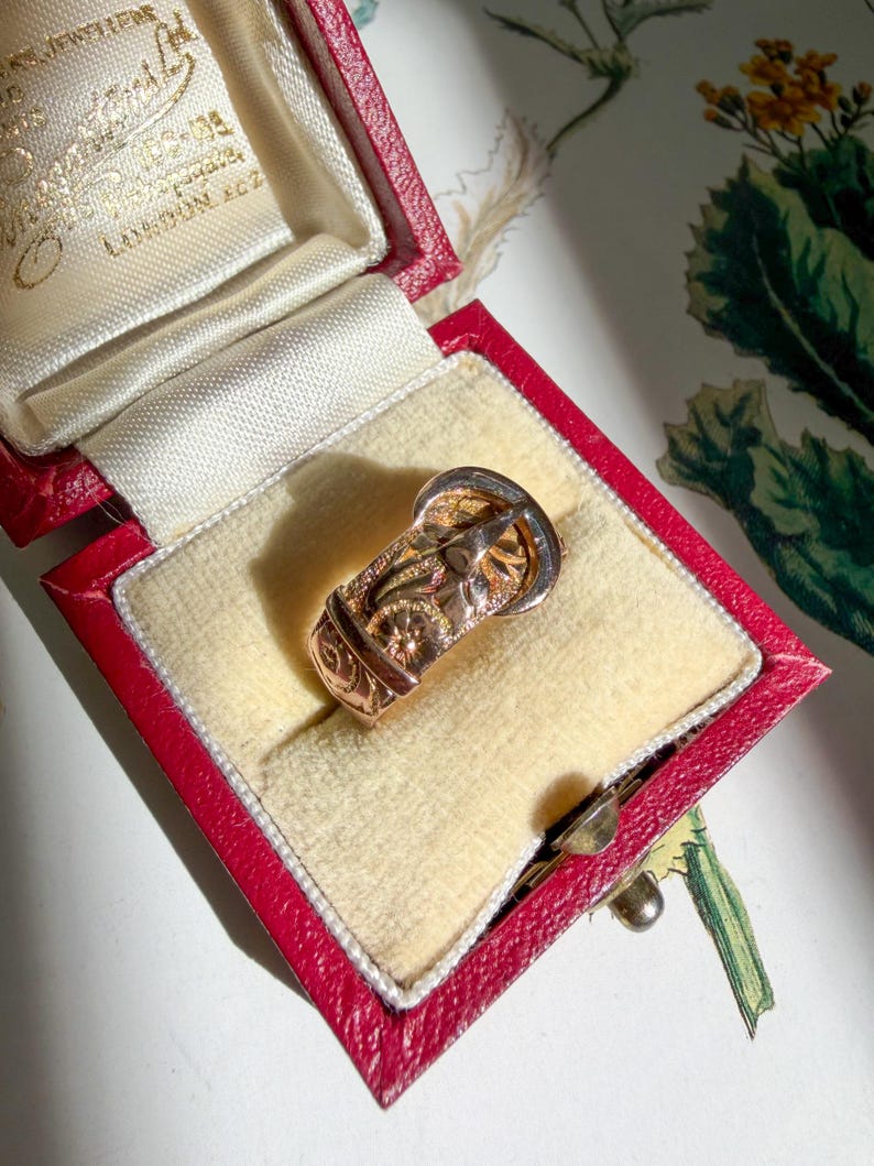 May include: A gold ring shaped like a belt buckle with engraved floral patterns and a textured finish, presented inside a red rectangular jewelry box with a cream-colored velvet interior lining and gold text printed on the inner lid.