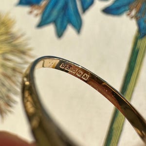 May include: Close-up of a gold ring with a hallmark stamp. The ring is set against a backdrop of blue and green floral patterns. The hallmark stamp is visible on the inside of the ring.