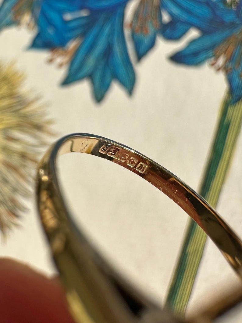 May include: Close-up of a gold ring with a hallmark stamp. The ring is set against a backdrop of blue and green floral patterns. The hallmark stamp is visible on the inside of the ring.