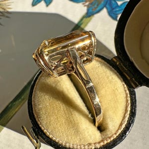 May include: A gold ring with a large, rectangular, amber-colored gemstone. The ring is set in a filigree design and is displayed in a black ring box with a yellow interior. The ring is shown against a floral background.
