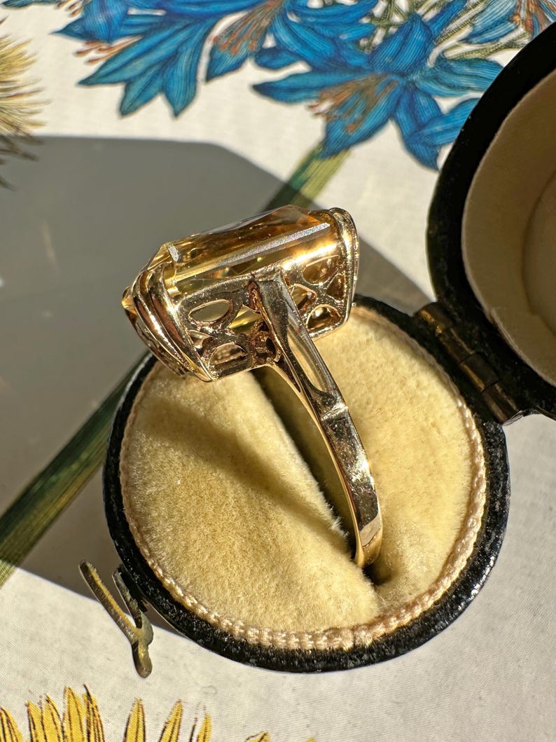 May include: A gold ring with a large, rectangular, amber-colored gemstone. The ring is set in a filigree design and is displayed in a black ring box with a yellow interior. The ring is shown against a floral background.