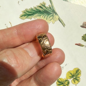 May include: A gold-toned band ring featuring an embossed floral pattern across its entire outer surface. The metal has a polished, reflective finish that catches the light, highlighting the intricate, raised botanical details carved into the metal.