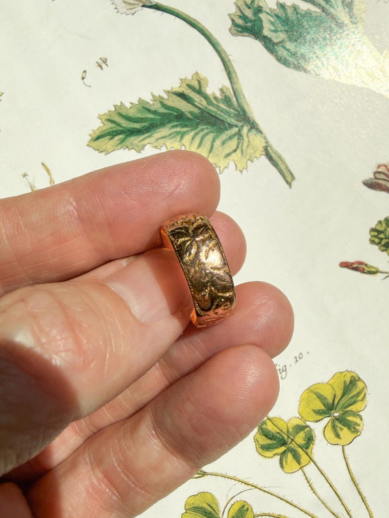 May include: A gold-toned band ring featuring an embossed floral pattern across its entire outer surface. The metal has a polished, reflective finish that catches the light, highlighting the intricate, raised botanical details carved into the metal.
