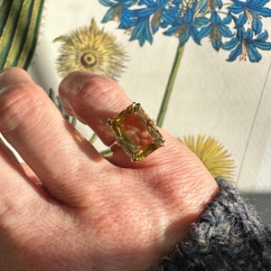 May include: A gold ring featuring a large, rectangular, amber-colored gemstone. The ring is set on a hand, with a floral illustration in the background. The gemstone is the focal point, catching the light and showcasing its color and cut.