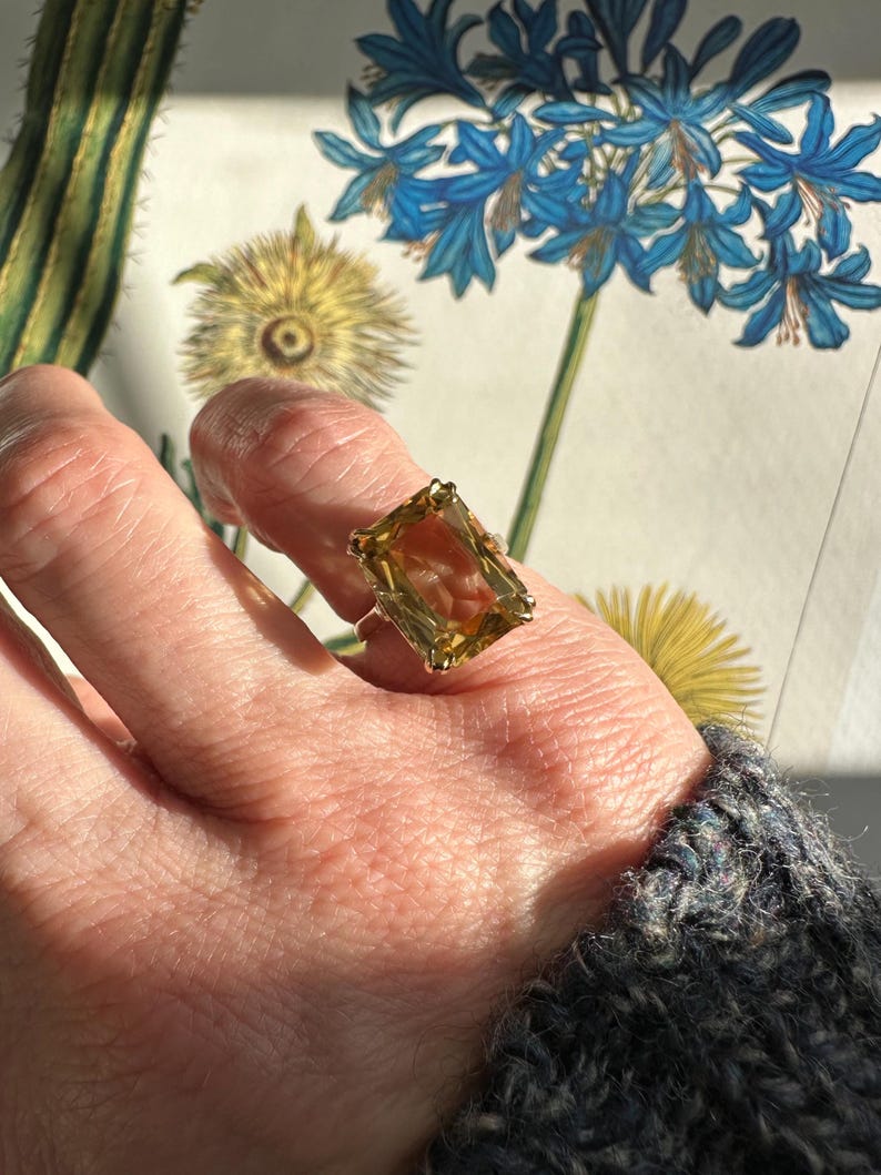 May include: A gold ring featuring a large, rectangular, amber-colored gemstone. The ring is set on a hand, with a floral illustration in the background. The gemstone is the focal point, catching the light and showcasing its color and cut.