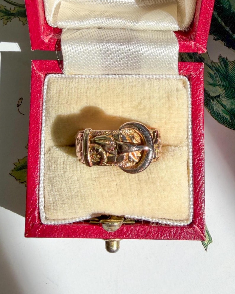 May include: A gold ring designed to resemble a miniature belt with a buckle, featuring an engraved pattern along the band. The ring is presented inside a small, square, red jewelry box with a cream-colored velvet interior lining.