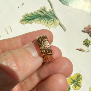 May include: A gold-toned band ring featuring an intricate, embossed floral pattern across its entire outer surface. The metal has a polished, reflective finish that catches the light, highlighting the detailed engravings of small flowers and leaves.