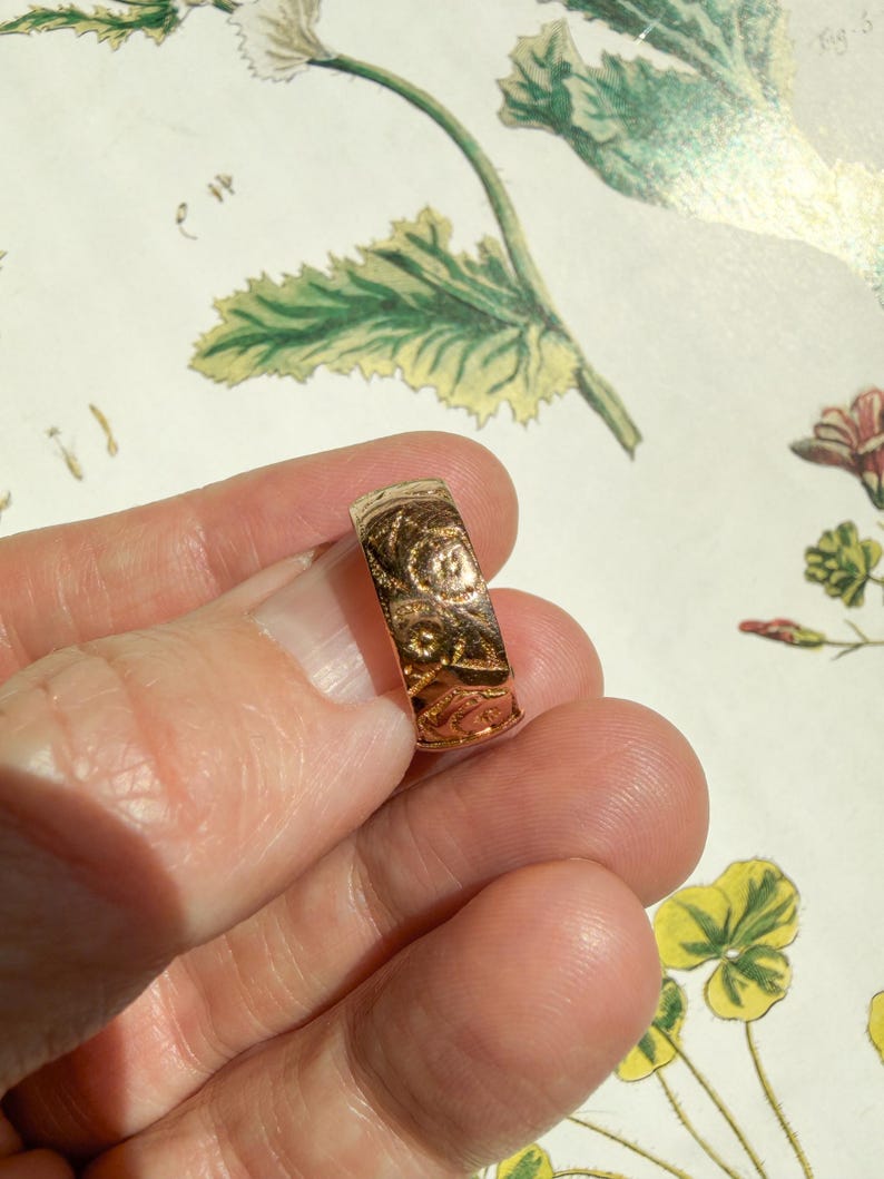 May include: A gold-toned band ring featuring an intricate, embossed floral pattern across its entire outer surface. The metal has a polished, reflective finish that catches the light, highlighting the detailed engravings of small flowers and leaves.