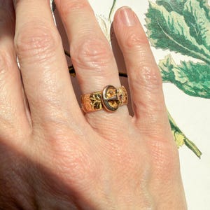 May include: A gold-toned metal ring featuring a buckle design. The band is decorated with an intricate, embossed floral pattern across its surface. The buckle component is positioned at the center, mimicking the appearance of a fastened belt.