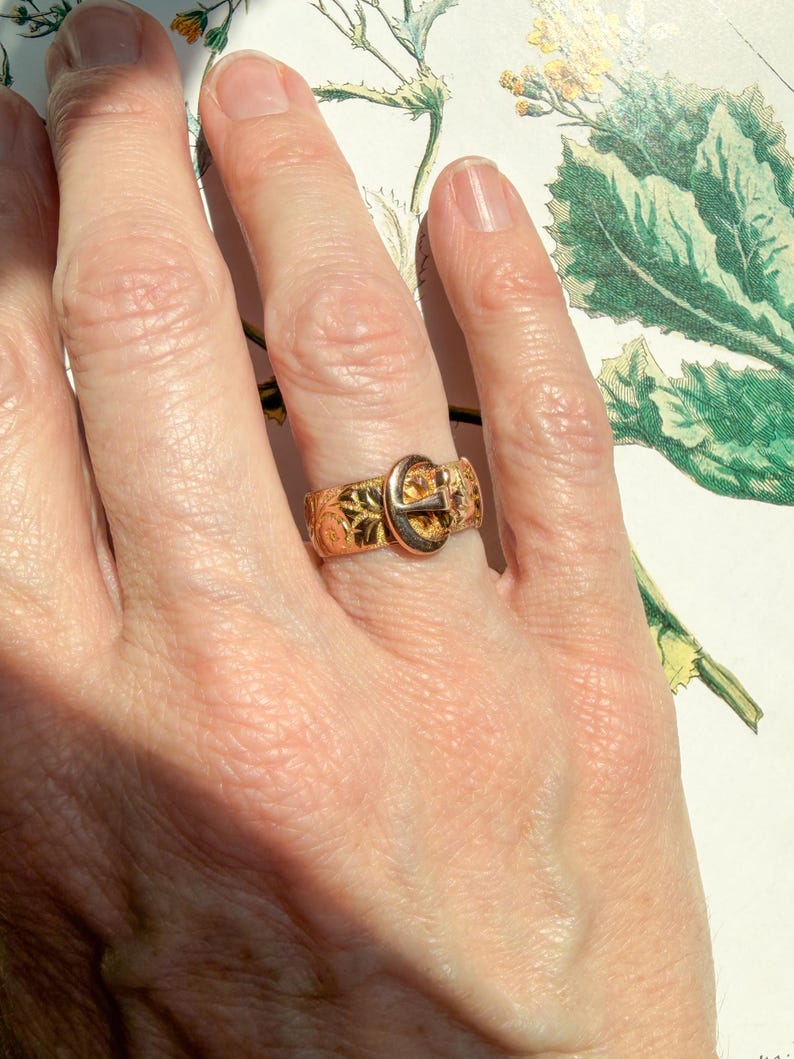 May include: A gold-toned metal ring featuring a buckle design. The band is decorated with an intricate, embossed floral pattern across its surface. The buckle component is positioned at the center, mimicking the appearance of a fastened belt.