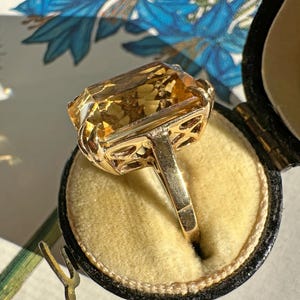 May include: A gold ring with a large, rectangular, amber-colored gemstone. The ring is set in a filigree design and is displayed in a black jewelry box with a yellow interior. The background features blue floral patterns.