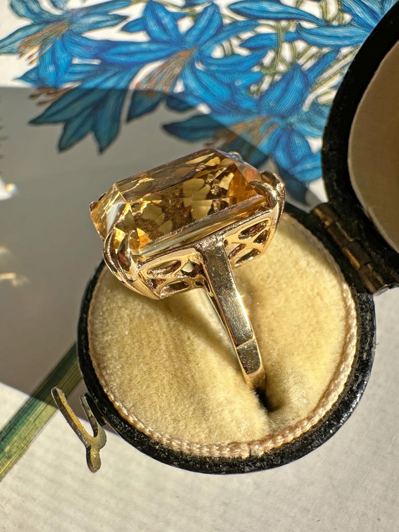May include: A gold ring with a large, rectangular, amber-colored gemstone. The ring is set in a filigree design and is displayed in a black jewelry box with a yellow interior. The background features blue floral patterns.