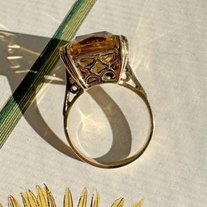 May include: A gold ring featuring a large, rectangular, amber-colored gemstone. The ring has an ornate, openwork design around the stone and on the band's shoulders. The ring is set against a floral patterned background.