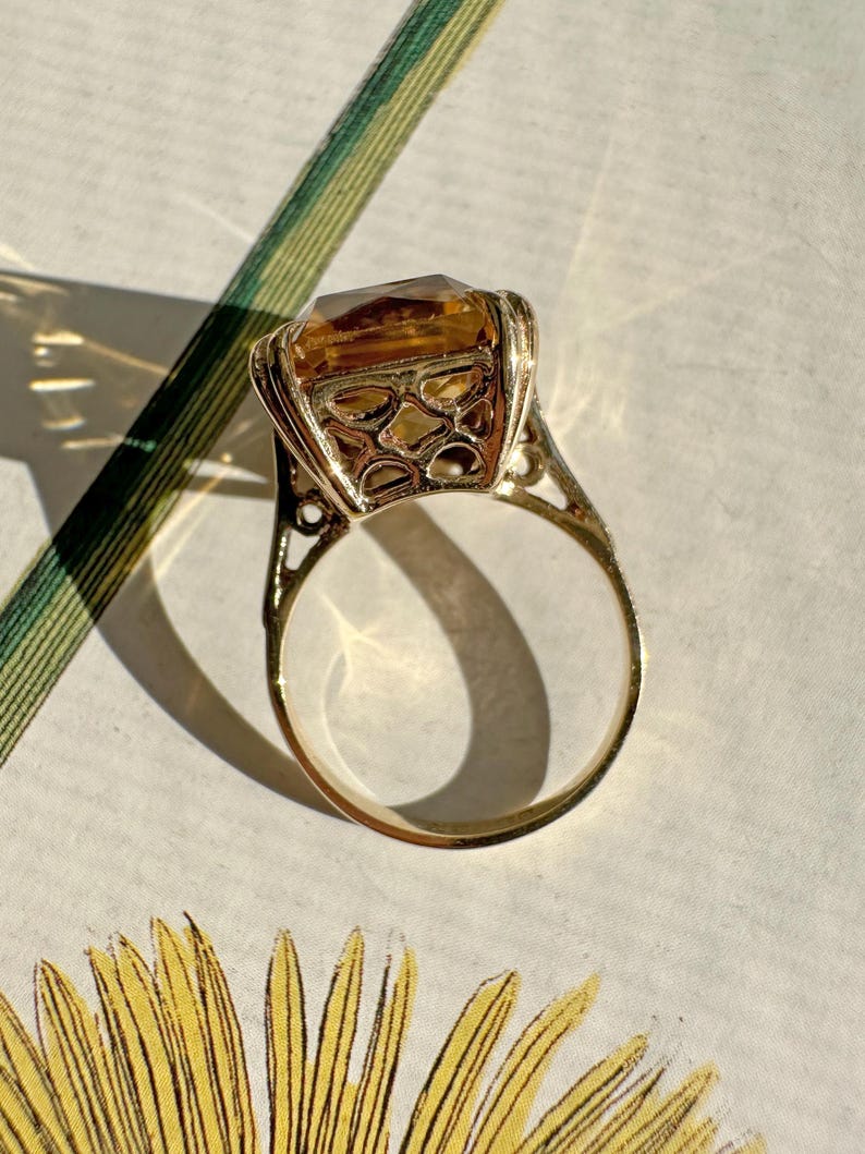May include: A gold ring featuring a large, rectangular, amber-colored gemstone. The ring has an ornate, openwork design around the stone and on the band's shoulders. The ring is set against a floral patterned background.