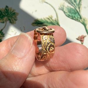 May include: A gold-toned ring featuring a buckle-shaped top and an intricate, embossed floral pattern covering the entire band. The metal surface displays a mix of yellow and rose gold hues with detailed engravings of leaves and small flowers.