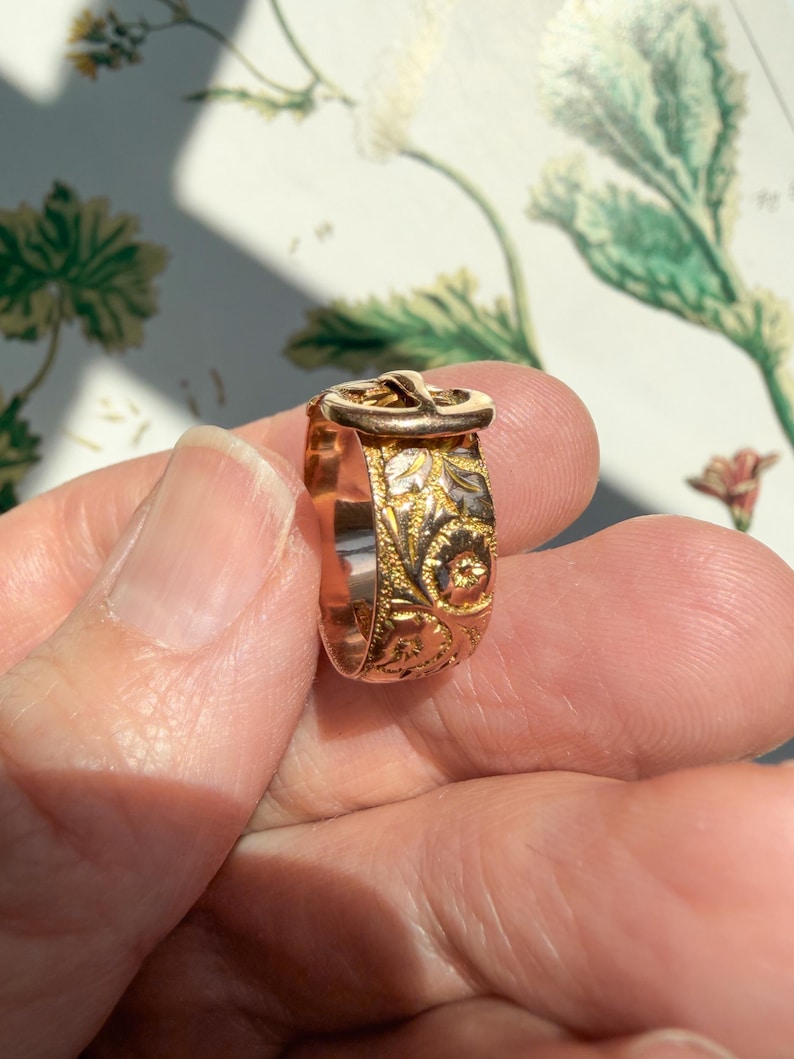 May include: A gold-toned ring featuring a buckle-shaped top and an intricate, embossed floral pattern covering the entire band. The metal surface displays a mix of yellow and rose gold hues with detailed engravings of leaves and small flowers.