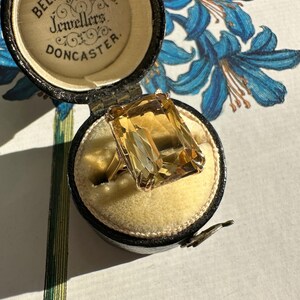 May include: A vintage gold ring with a large, rectangular, amber-colored gemstone, displayed in an open black jewelry box with the text "Bell Brothers Jewellers Doncaster". The box has a yellow velvet interior.