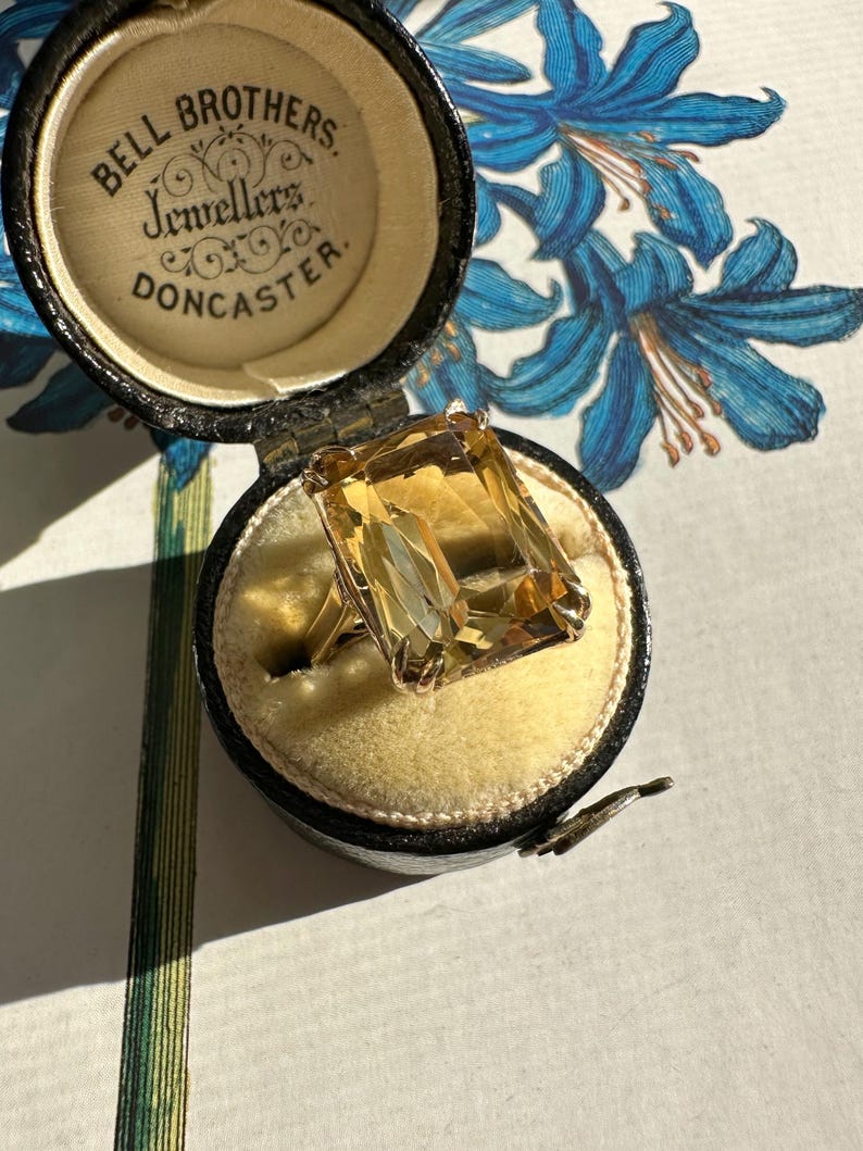 May include: A vintage gold ring with a large, rectangular, amber-colored gemstone, displayed in an open black jewelry box with the text "Bell Brothers Jewellers Doncaster". The box has a yellow velvet interior.