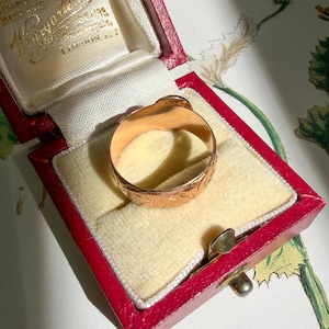 May include: A gold ring with an engraved floral pattern on the outer band, resting inside a red rectangular jewelry box with a cream-colored velvet interior. The box lid features gold embossed text that reads "Viney & Sons Ltd".